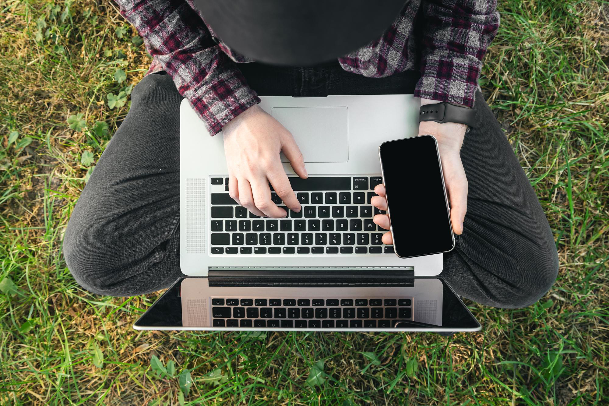 man-sits-grass-with-laptop-smartphone-top-view